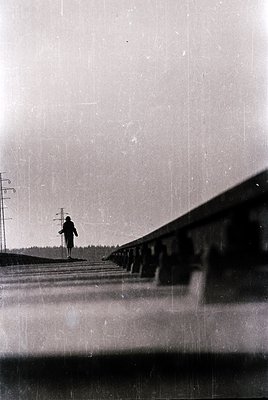Vintage black-and-white photograph of a lone figure walking alongside a railway track, framed by a low concrete barrier. Over...
