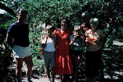 Family portrait in lush greenery, mid-20th century. Five individuals pose outdoors, holding watermelons. Adults wear patterne...