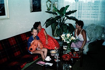 Vintage indoor scene featuring three people in a cozy 1970s living room. A woman in a red floral dress sits beside a boy in a...