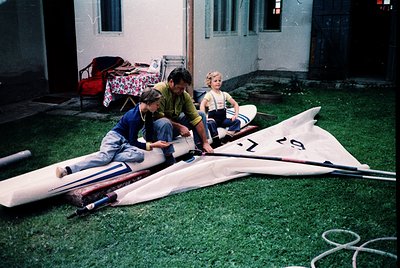 Three children assemble a sailboat in a grassy courtyard, 1970s. White sail with red "19" marking and blue hull on light gree...