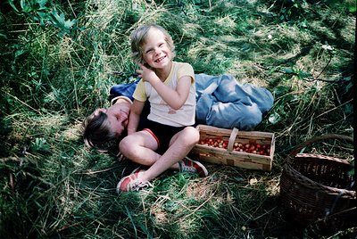 Two children rest in tall grass, surrounded by harvested cherries in wooden and woven baskets. The adult’s vintage clothing (...