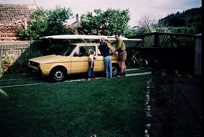 Family poses beside a vintage **Škoda 120L** station wagon (1970s) with surfboard rack, suggesting coastal or recreational tr...
