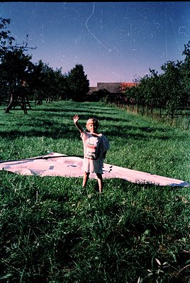 Vintage photo of a child standing on a pink inflatable mat in a lush, tree-lined field under clear skies. Mid-20th century ou...
