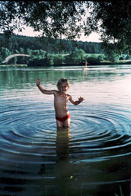 A young child joyfully splashes in shallow, reflective water under a leafy tree, surrounded by lush greenery and a distant br...