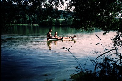 Two individuals relax in a wooden rowboat on calm waters surrounded by lush greenery, likely during mid-20th century. Warm go...