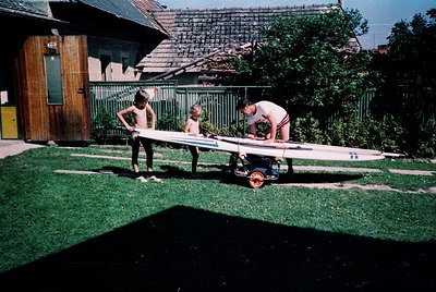 A man in swim trunks and a striped shirt pushes a vintage surfboard on a cart, accompanied by two children in shorts, likely ...