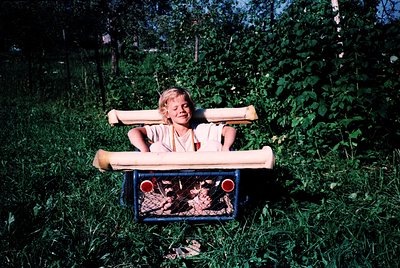 Vintage childhood joy: child riding homemade wooden cart through lush greenery, mid-20th century. Bright red wheels contrast ...
