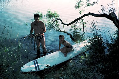 Two individuals prepare for paddleboarding in a serene lake setting, surrounded by dense reeds. The man stands holding a padd...