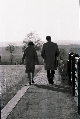 Couple walking away on a paved riverside path, dressed in 1960s formal attire—man in overcoat, woman in knee-length skirt. Ov...
