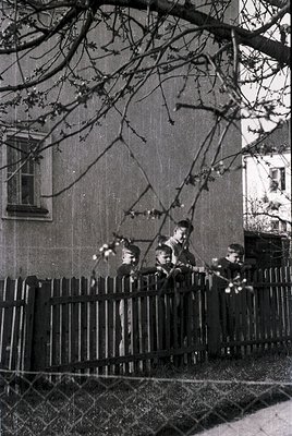 Four boys in school uniforms lean over a black wrought-iron fence, tossing white paper balls under bare winter trees. Mid-20t...