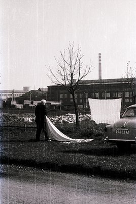 Mid-20th century industrial scene: man in suit unfurls a large white banner near a vintage car. Factory with smokestack and b...