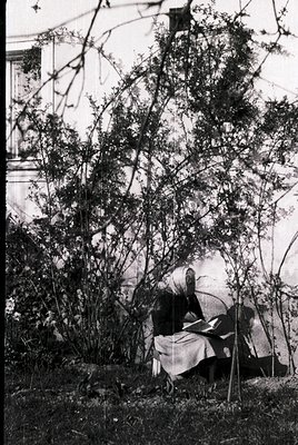 Vintage black-and-white photo of two seated figures in a garden, partially obscured by leafless branches. One figure holds a ...