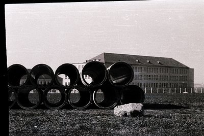 Large concrete pipes arranged in a row, partially obscuring a multi-story institutional building with gabled roofs. Mid-20th ...