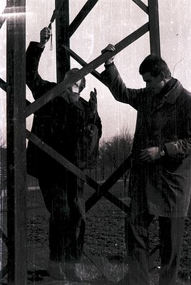Black-and-white photo of three men constructing a metal framework, likely a tower or structure, in an outdoor setting. Clothi...