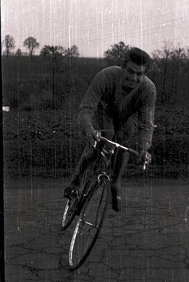 Black-and-white shot of a cyclist mid-motion on a roadside, likely 1960s–1970s. The rider wears a sleeveless jersey and helme...