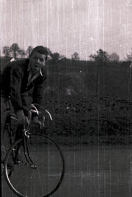 Vintage black-and-white photo of a man riding a classic road bike, likely mid-20th century. His patterned suit and helmet sug...