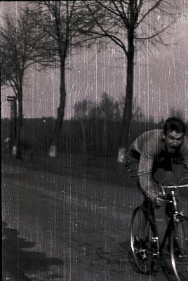 Vintage black-and-white photo of a cyclist riding through rain on a rural road, surrounded by bare trees. The rider wears a l...