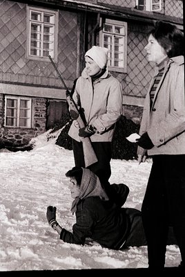 Cold winter scene featuring three individuals in snowy courtyard. Two adults in winter coats and hats stand beside a child kn...