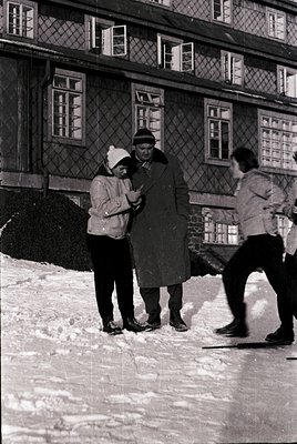 Three individuals in winter attire stand on a snow-covered surface beside a wooden building with diamond-patterned window pan...