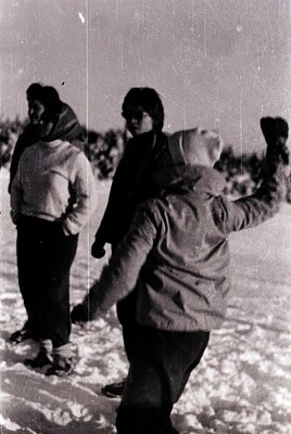 Vintage black-and-white photo of three individuals playing snowballs in an open, snowy field. Mid-20th century winter attire:...