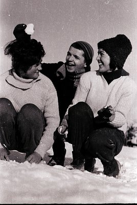 Three individuals in winter attire pose joyfully on snow-covered ground, mid-laughter. Knit sweaters, beanies, and gloves sug...