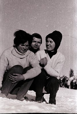 Black-and-white snapshot of three individuals posing on snow-covered ground, likely a ski resort. The man sits centrally in a...