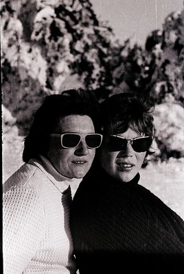 Two individuals pose outdoors in 1960s-style sunglasses and casual attire, likely mid-20th century. The woman on the left wea...