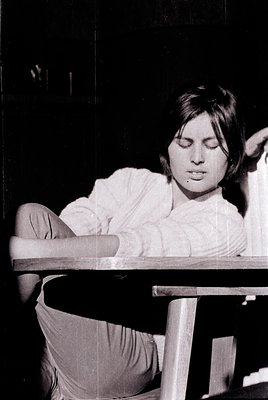 Black-and-white portrait of a person resting their head on a wooden table, seated in a chair. Lighting emphasizes soft shadow...