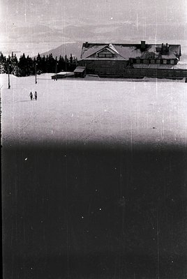 Vintage black-and-white ski lodge set against snow-covered slopes, likely mid-20th century. Two skiers in classic attire desc...