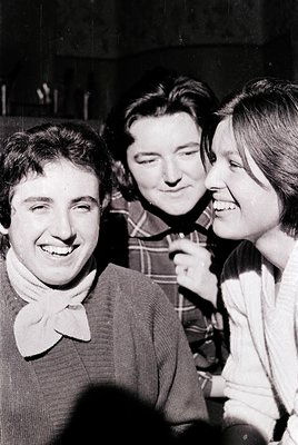 Three individuals in casual 1970s attire—men’s plaid shirt, collared sweater, and woman’s blouse—pose indoors with warm smile...