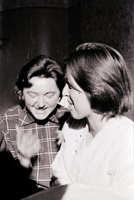 Black-and-white candid of two women sharing a playful moment, likely mid-1960s–70s. Plaid shirt and blouse with a choker neck...