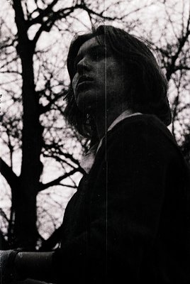 Portrait of a person in profile, backlit by bare trees, suggesting late autumn/winter. Long hair, high-collared coat, and a c...