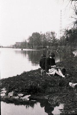 Two individuals sit on rocky riverbank, engrossed in conversation or reading. Mid-20th century clothing suggests or . Industr...