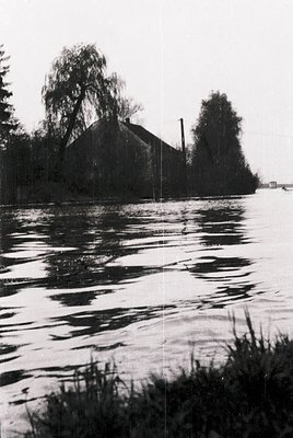 Black-and-white lakeside scene featuring a weathered, single-story building with a sloped roof and chimney, partially obscure...