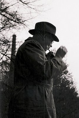 Mid-20th century man in outdoor setting, adjusting a tie while holding a hat. Dark coat, fedora, and barber-style apron sugge...