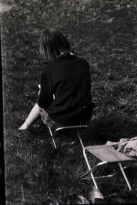 A person sits alone on a folding chair in tall grass, facing away, wearing a dark sweater and checkered socks. The vintage mo...