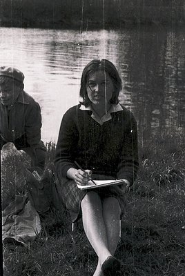 Black-and-white photo of a woman in mid-20th century attire (1950s–60s) sketching by a serene lake, seated on grass. Her swea...