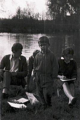 Three individuals by a lake, mid-20th century. Left: seated woman writing in notebook, holding a folded paper. Center: standi...