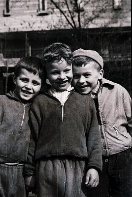 Three boys in early 20th-century streetwear pose joyfully outdoors, likely mid-1900s. Wearing button-up shirts, collars, and ...