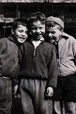Three boys in early-to-mid 20th-century streetwear pose closely together, radiating joy. Left wears a long-sleeve shirt with ...