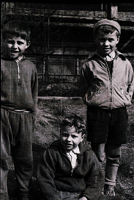 Three boys in mid-20th-century streetwear pose outdoors near scaffolding and brick ruins. Left: boy in striped sweater and pa...