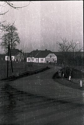Vintage black-and-white street scene featuring a row of mid-20th century residential buildings with gabled roofs and white fa...