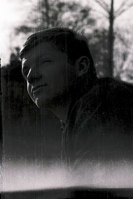 Portrait of a man in mid-smile, captured through rain-streaked glass. His short hair and light-colored shirt suggest a mid-20...
