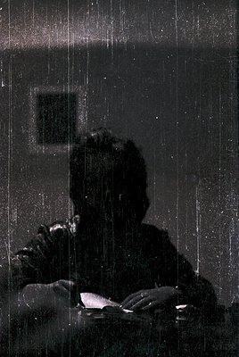Silhouetted figure reading indoors under heavy rain, captured through a frosted glass window. Classic monochrome composition ...