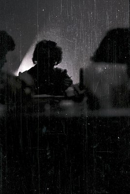 Silhouetted figure seated indoors, framed by rain-streaked window glass. Mid-20th century interior with visible curtains and ...