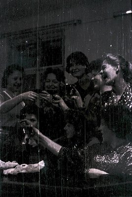 Vintage black-and-white photo of a lively group of five people gathered indoors, likely in the 1960s-70s. A man in a dark shi...