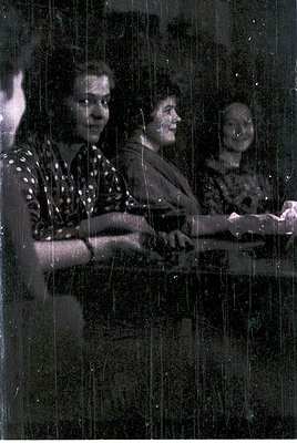 Black-and-white photo of three seated women in a dimly lit indoor setting, possibly a café or lounge. The woman on the left w...
