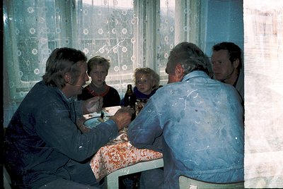 Family gathering in a modest indoor setting, likely 1970s–1980s. Five adults seated around a table with a floral tablecloth, ...