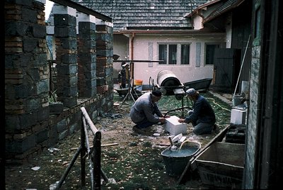 Two men craft traditional pottery in a courtyard using hand tools and clay. Stone chimney stacks and a rustic wooden roof fra...