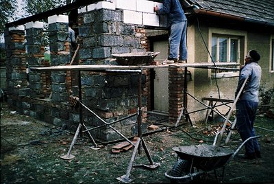 Three construction workers renovate a brick corner of a residential building, using scaffolding and cement mixers. The struct...